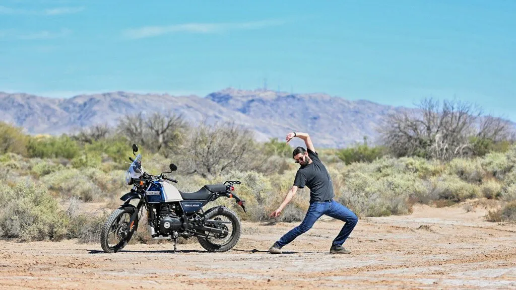 A black motorcycle is parked on a dirt road between two men, who stand against a backdrop of dry scrub brush and hazy mountains under a pale blue sky.