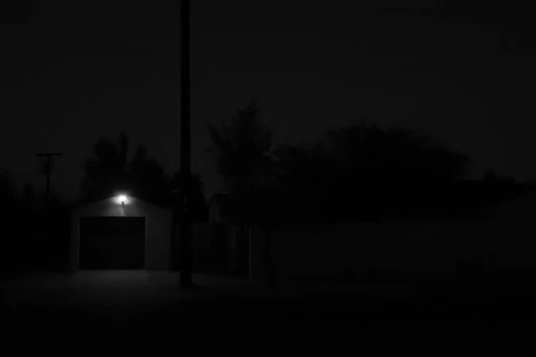 The composition is dominated by deep shadows of trees and a garage entrance, with a single, bright, warm light source visible above the door.