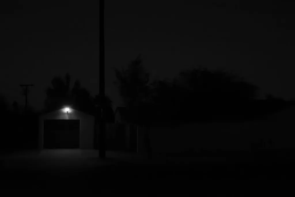 The composition is dominated by deep shadows of trees and a garage entrance, with a single, bright, warm light source visible above the door.
