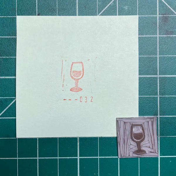 On a dark teal grid, a white paper displays a red line drawing of a wine glass and the number --032, positioned above two small, square artworks, one of which shows a textured reddish-brown wine glass.