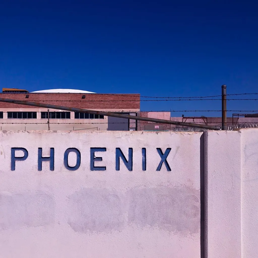 Large dark letters spelling PHOENIX are painted on a textured light pink wall, set against a backdrop of a reddish building and a clear blue sky.