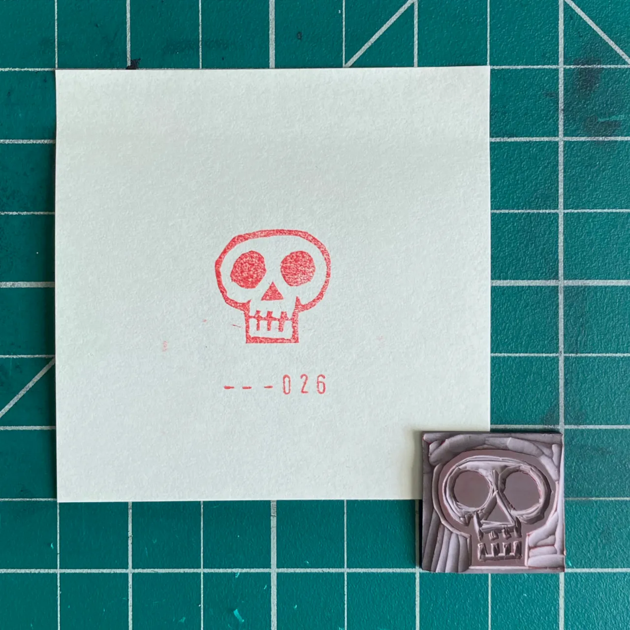 On a teal grid background, a white paper features a red outline drawing of a skull and the number --026, next to a small, molded pink skull object.