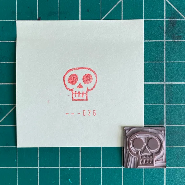 On a teal grid background, a white paper features a red outline drawing of a skull and the number --026, next to a small, molded pink skull object.