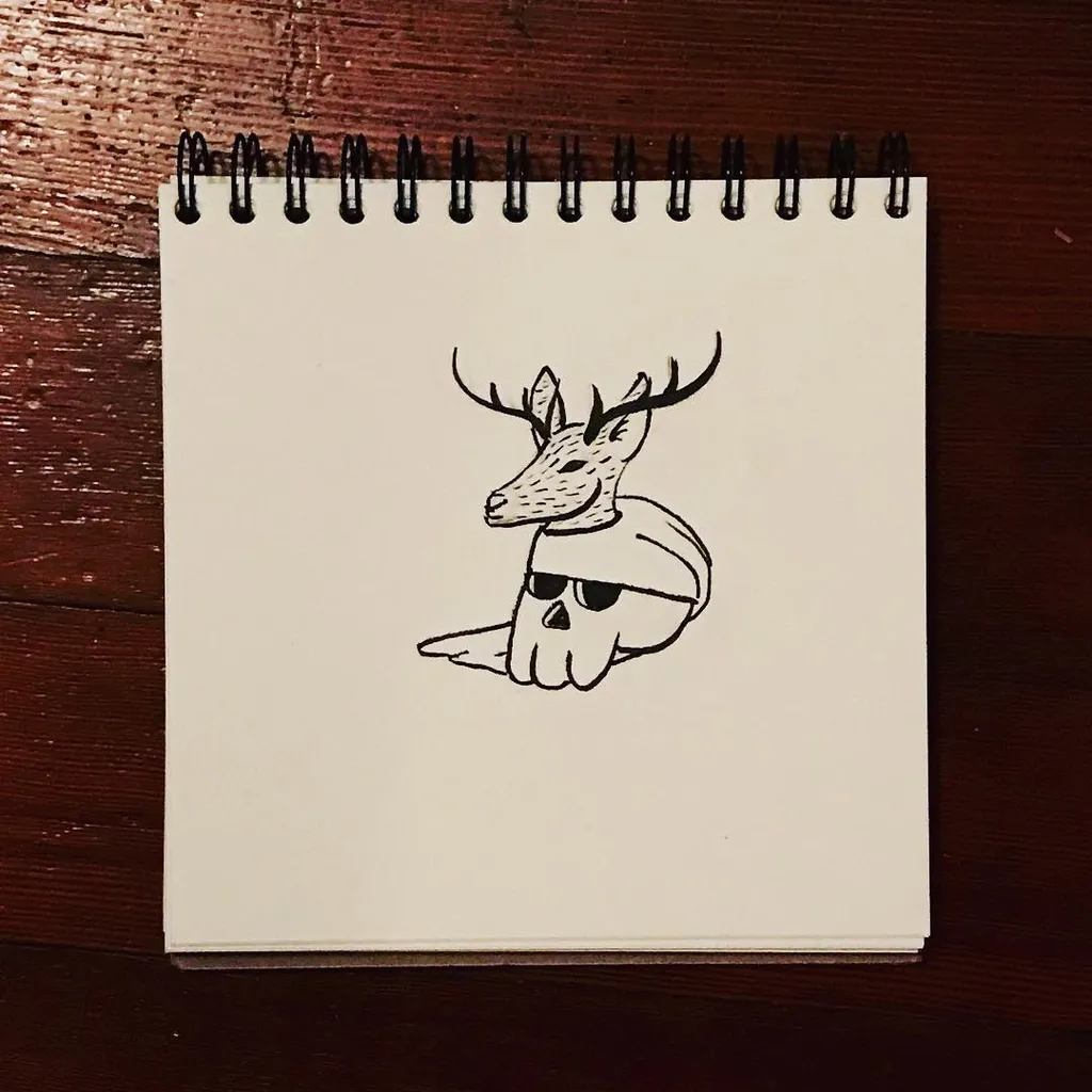 A black ink line drawing of a deer head wearing sunglasses and a bandana, centered on a spiral-bound notepad on a dark wooden surface.