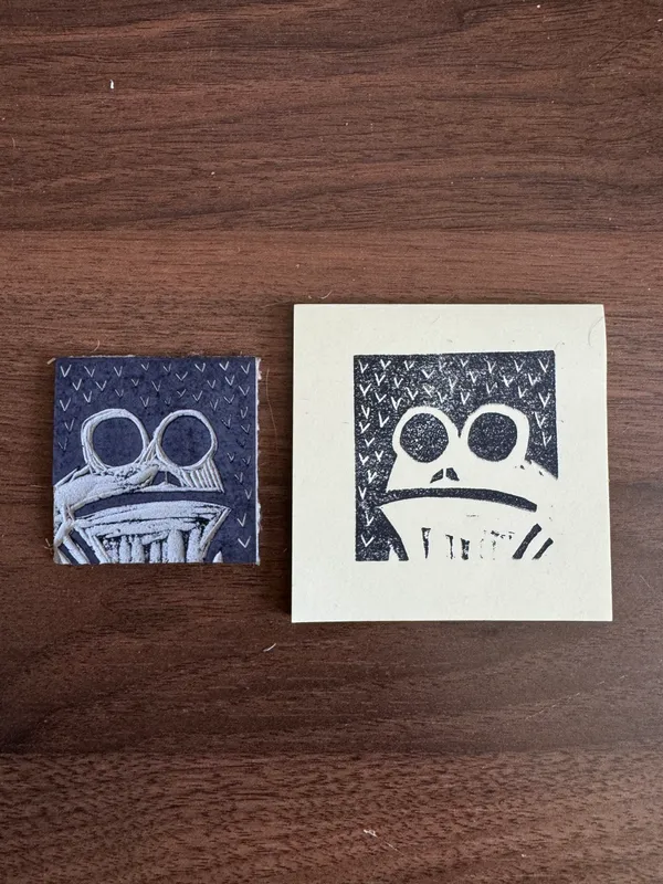 Two graphic, square prints rest on dark wood. The left print uses white ink on a dark background, depicting a patterned face; the right print uses black ink on white paper, showing a stylized face with large eyes.
