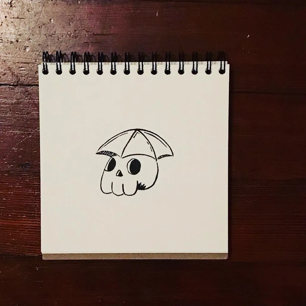 A simple black line drawing of a skull wearing an open umbrella is centered on a cream-colored spiral notebook page, which rests on a dark wooden surface.