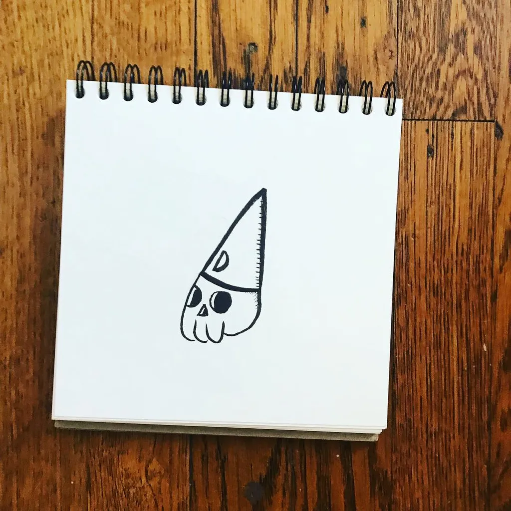 A simple black line drawing of a triangular skull face is placed on a white spiral-bound notepad, which sits on a dark, textured wooden surface.