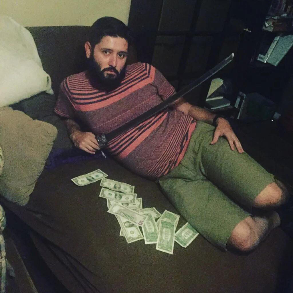 A bearded man in a striped shirt and green shorts sits on a dark surface, resting a sword across his lap above scattered dollar bills.