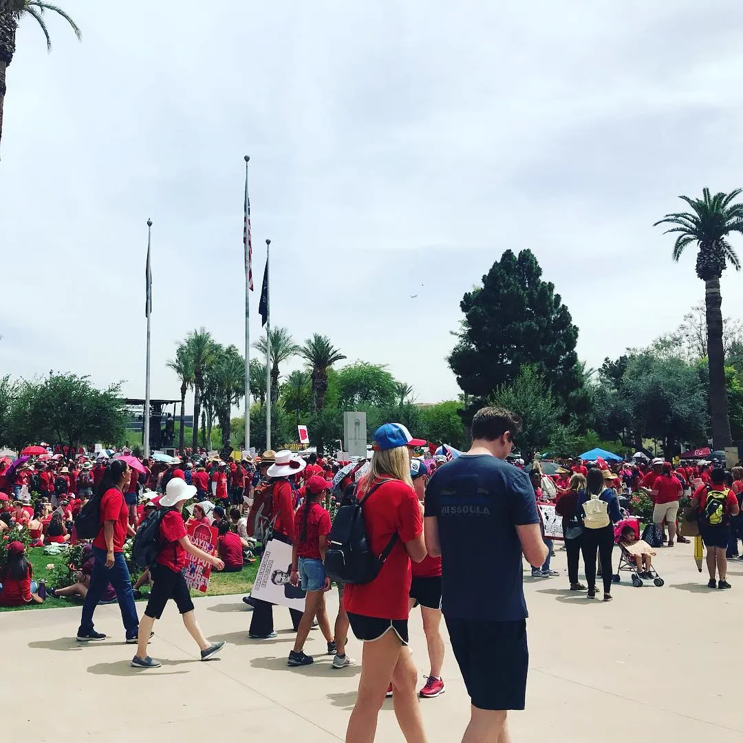 A large crowd of people gathers in a sunny outdoor plaza, with several individuals in the foreground standing on pavement beneath tall palm trees and a cloudy sky.