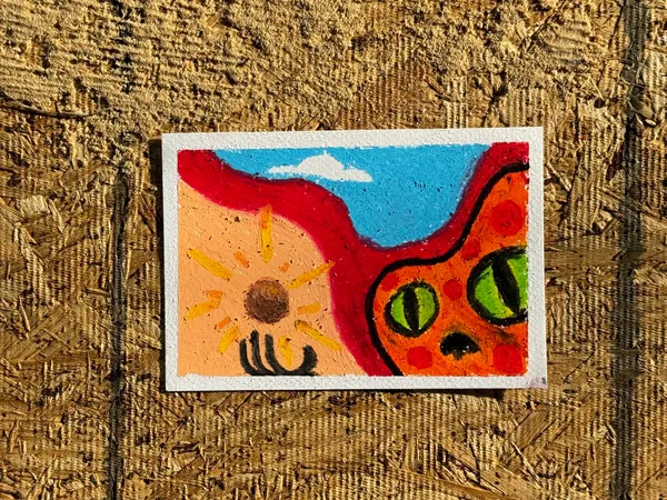 A stylized face with large green eyes and an orange body is painted on a canvas, featuring a yellow sun on the left and a blue sky above.