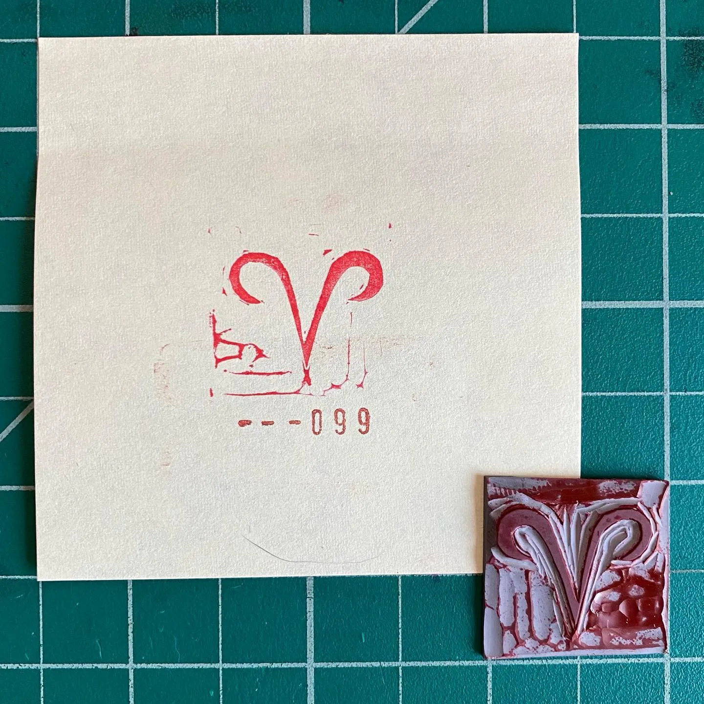 On a green grid, a cream card bears red markings, including a stylized V and numbers, next to a small, square object featuring abstract swirls of red, white, and gray.
