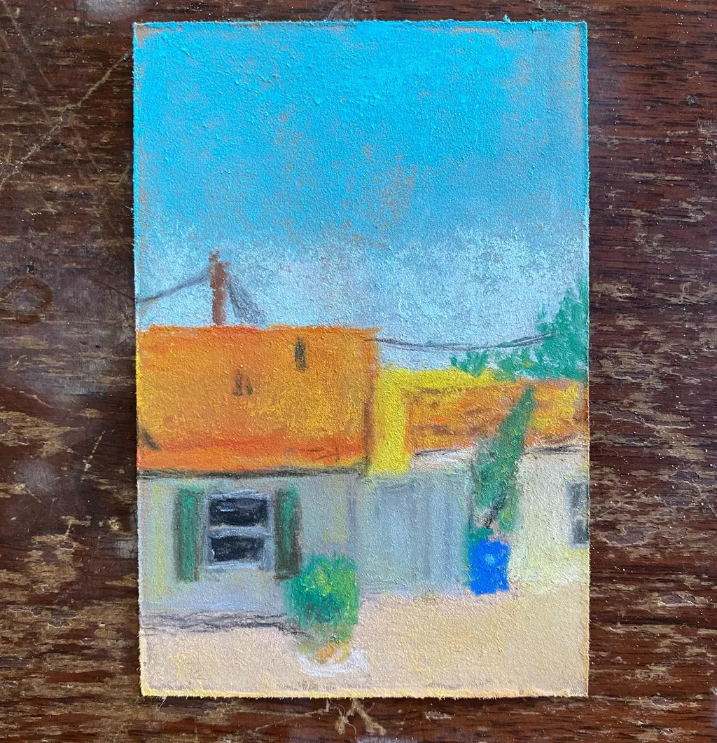 Bright blue sky dominates the upper half of the composition, above clustered buildings featuring orange roofs, white walls, and accents of green and blue, all painted on a dark wood surface.