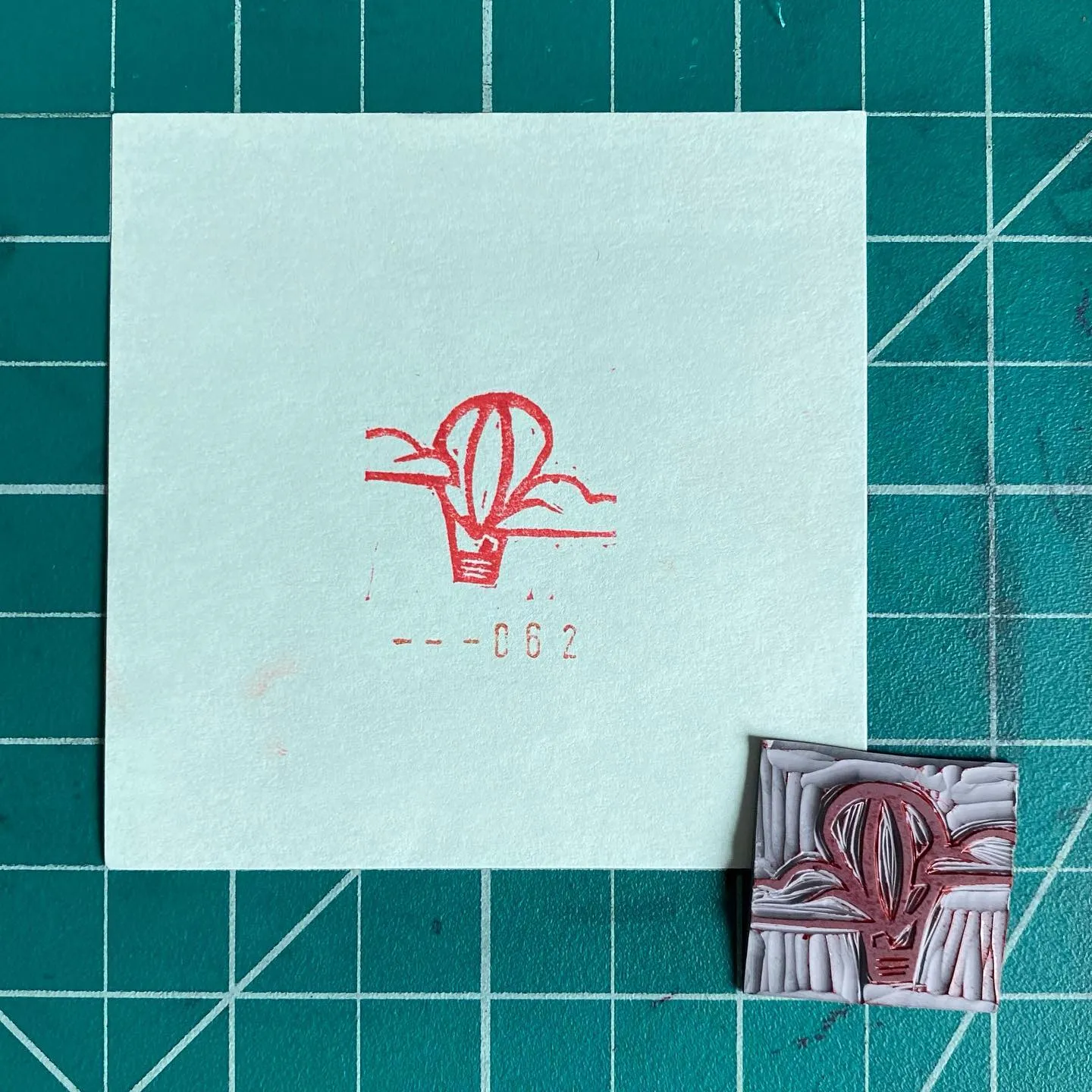 On a teal grid, a white paper displays a red line drawing of a potted plant and the number --062, next to a small, square, textured relief piece in red and white.