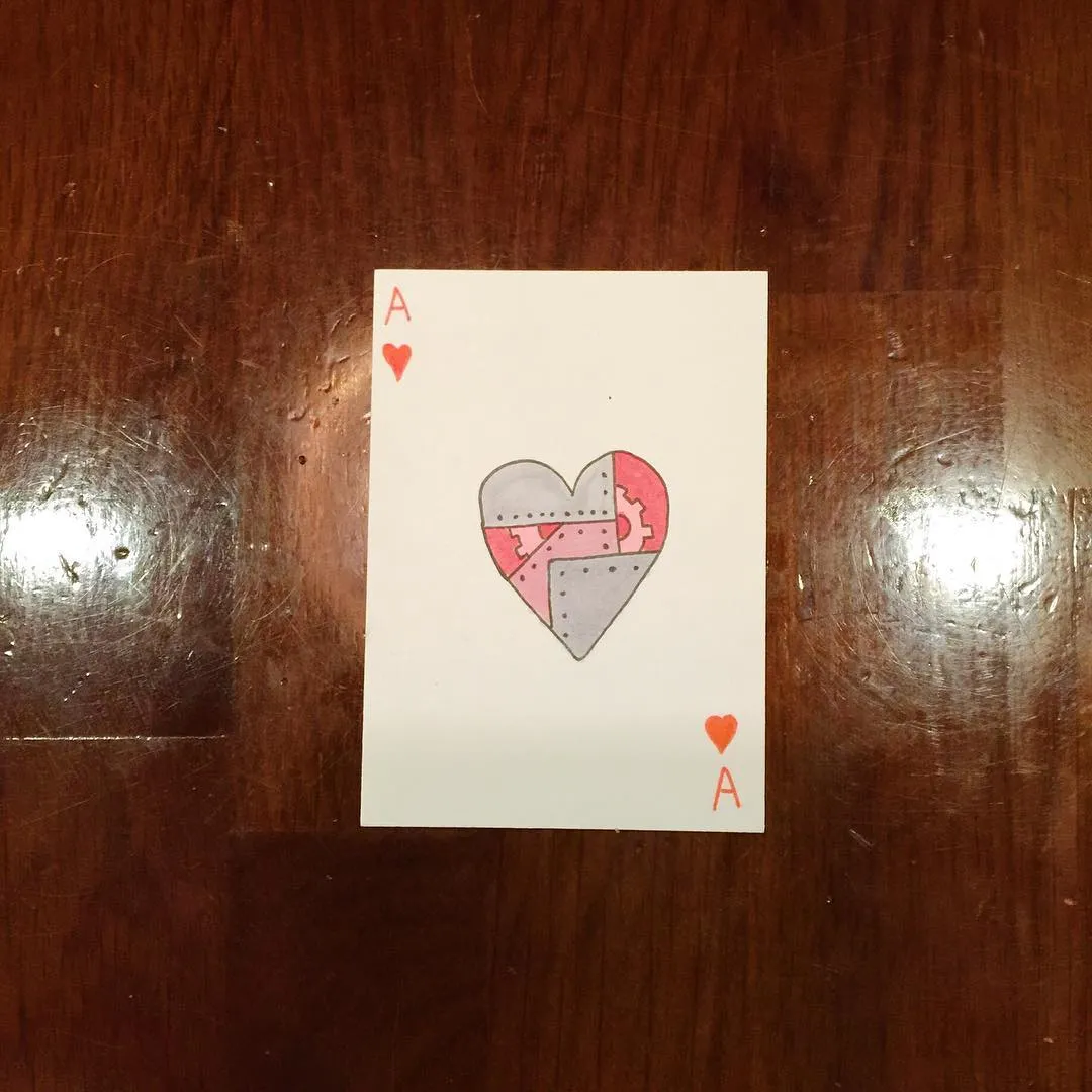 The card rests on dark wood grain, featuring the Ace of Hearts and a central drawing of a heart composed of three interlocking puzzle pieces in red and gray tones.
