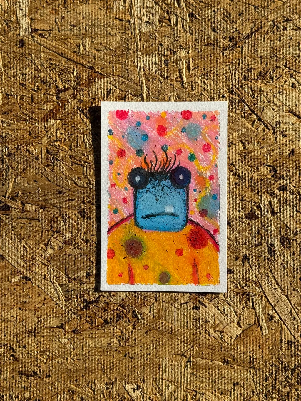 A stylized yellow creature with dark eyes and antennae is painted against a speckled pink and blue background, resting on a textured, brown plywood surface.