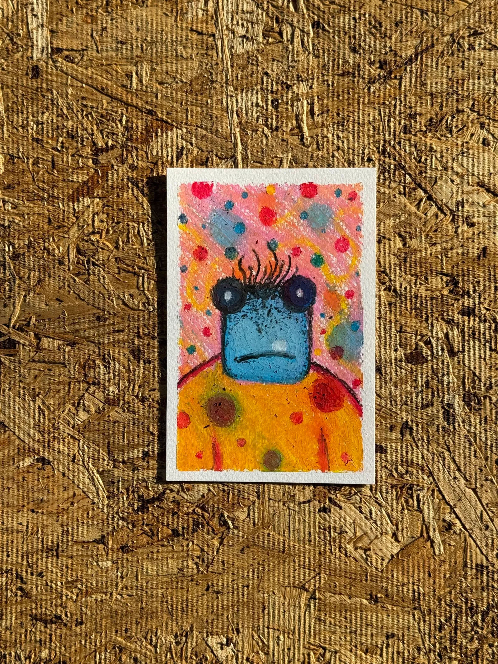 A stylized yellow creature with dark eyes and antennae is painted against a speckled pink and blue background, resting on a textured, brown plywood surface.