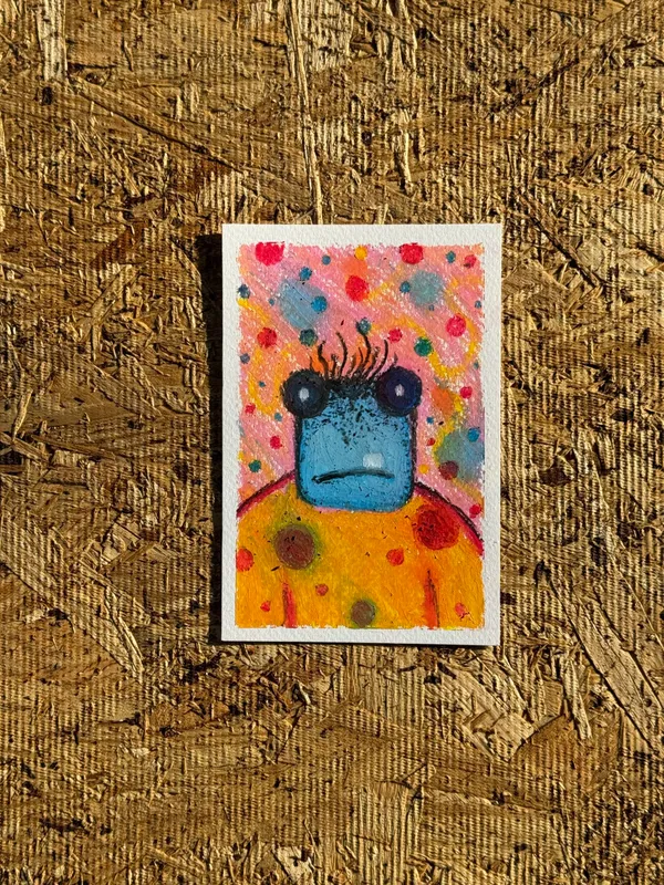 A stylized yellow creature with dark eyes and antennae is painted against a speckled pink and blue background, resting on a textured, brown plywood surface.