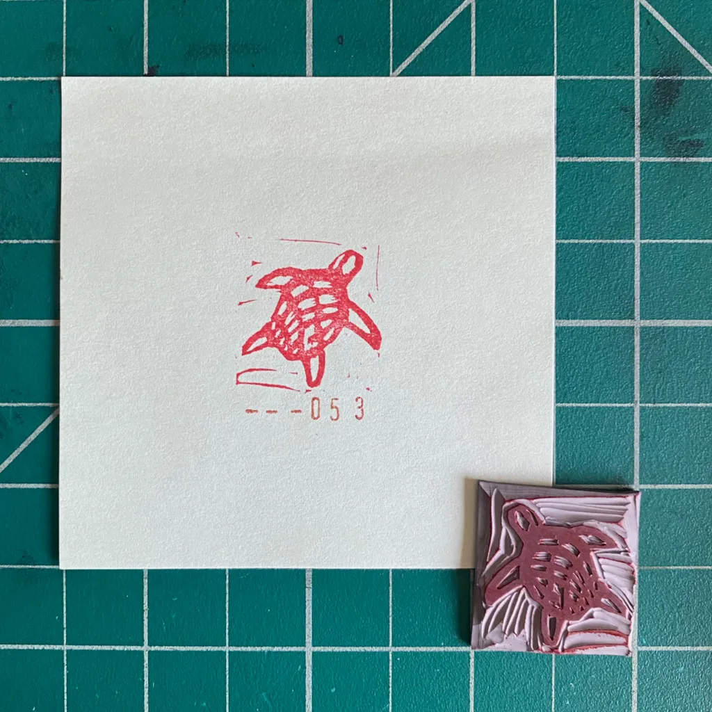 A white card on a teal grid displays a red outline sketch of a sea turtle with the numbers --053 below it, next to a small, pressed reddish-brown sea turtle shape.