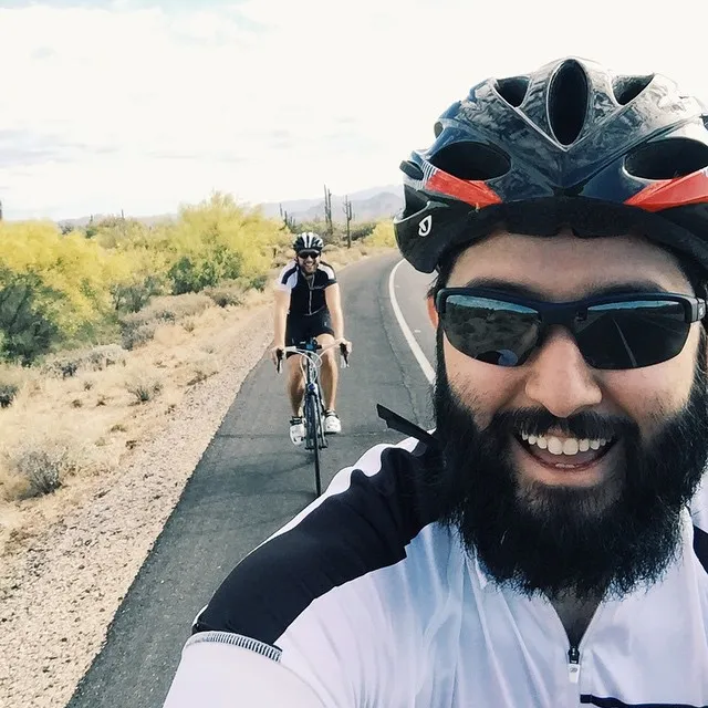 A bearded man in a cycling helmet smiles into the camera, positioned on the right side of a paved road that leads through dry, scrubby landscape where another cyclist rides in the distance.