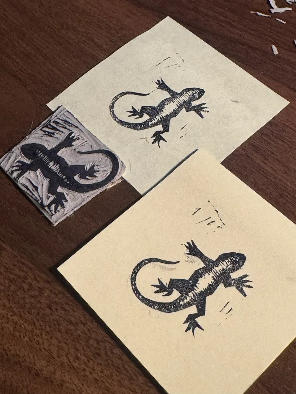 Three black ink sketches of lizards are displayed on a dark wooden surface, featuring varying styles: a detailed square drawing on the left, and two lighter sketches in the center.