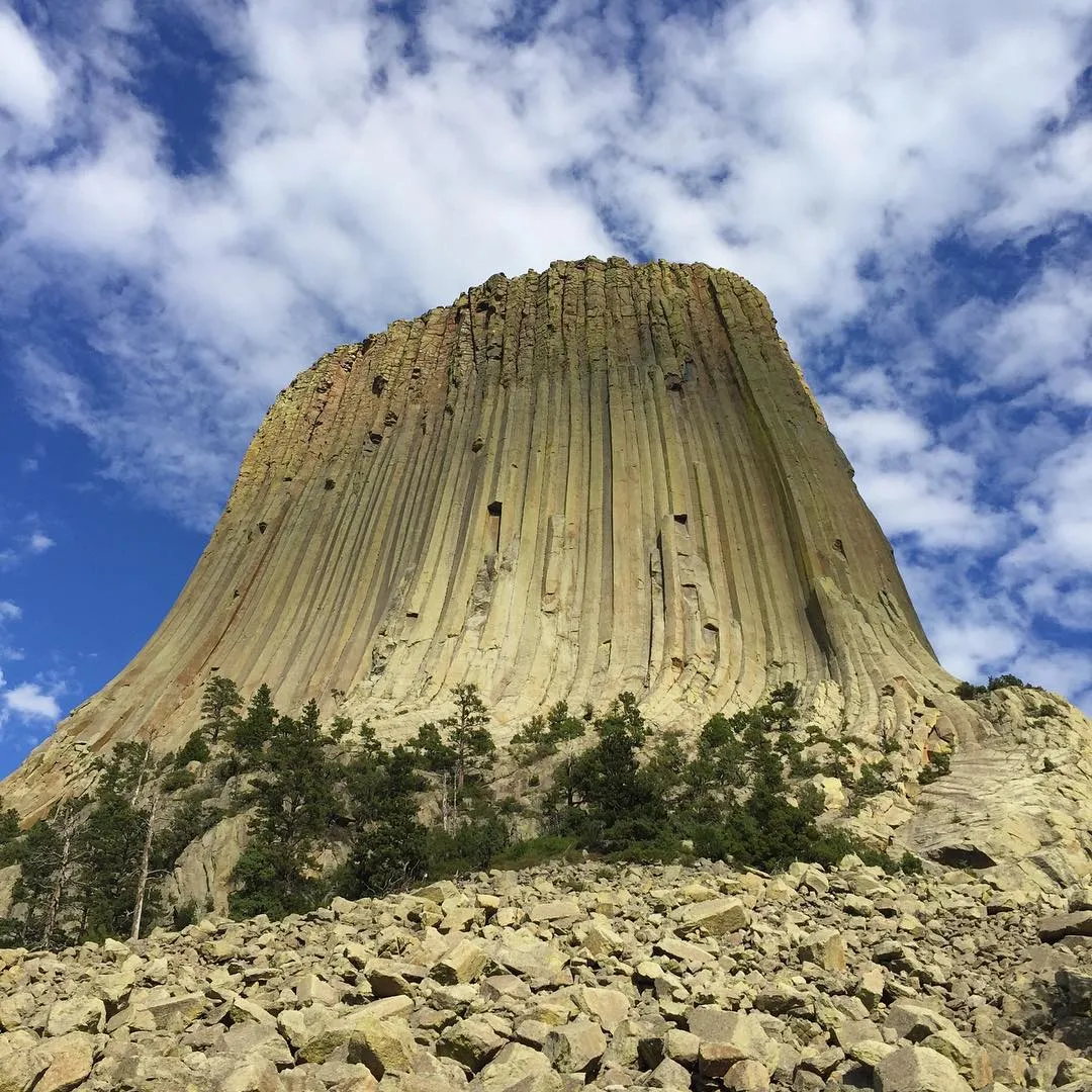 A massive, striped tan geological formation rises sharply against a blue sky with white clouds, viewed from a foreground of scattered gray rocks and green scrub.