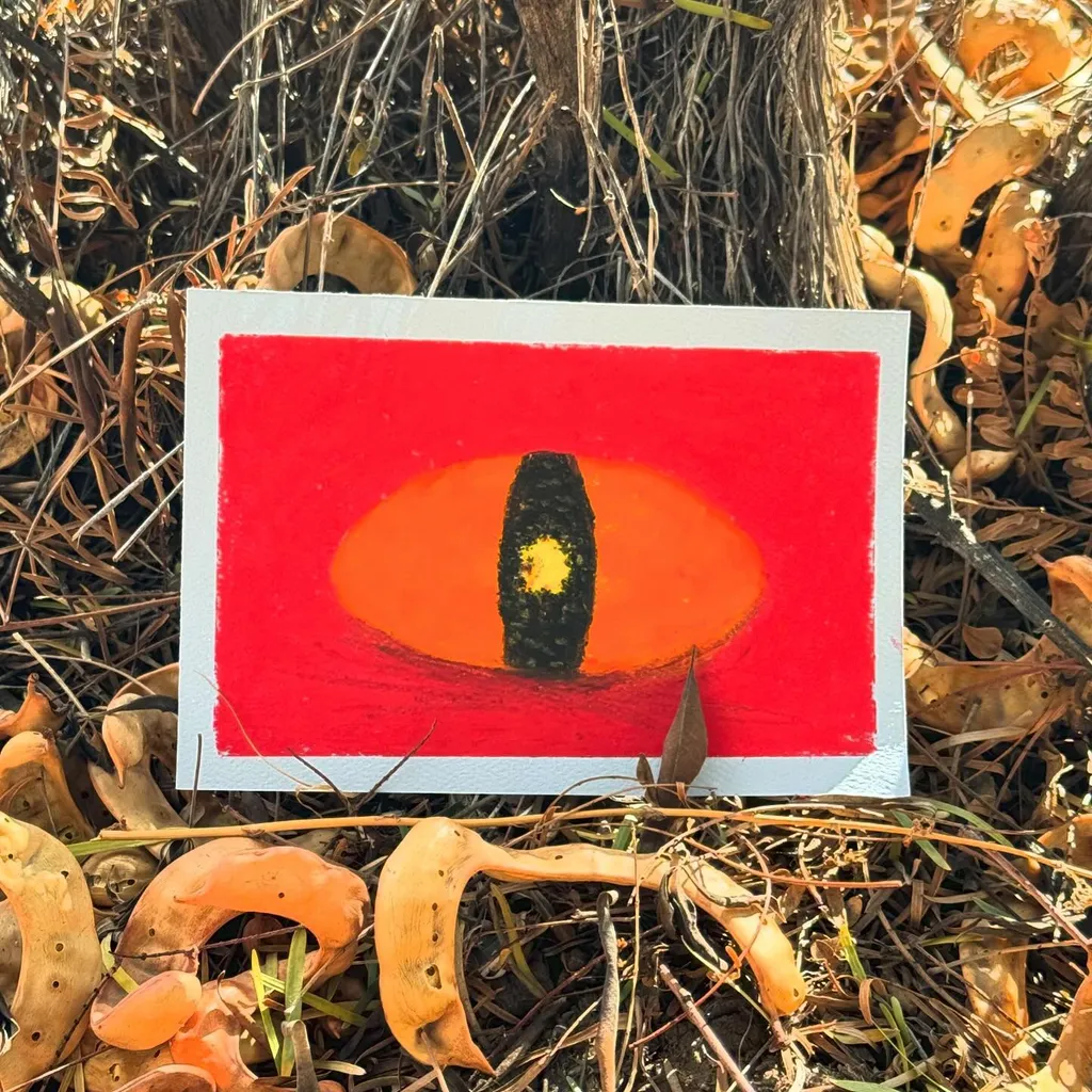 A square artwork with a red border, displaying an orange semi-circle and a central black oval marked with a yellow dot, rests on a bed of dry leaves and mushrooms.