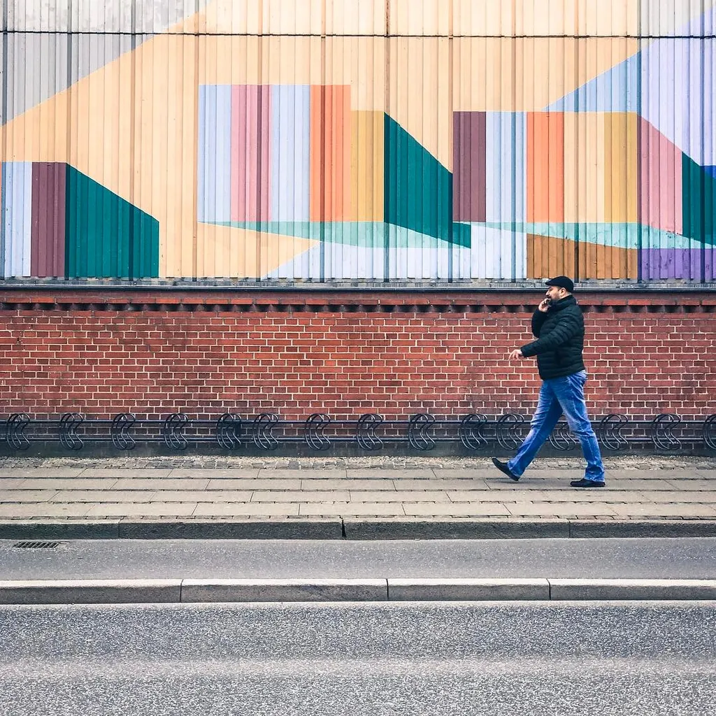 A man walks past a red brick wall and a large mural featuring abstract pastel vertical stripes and geometric shapes on a light background.