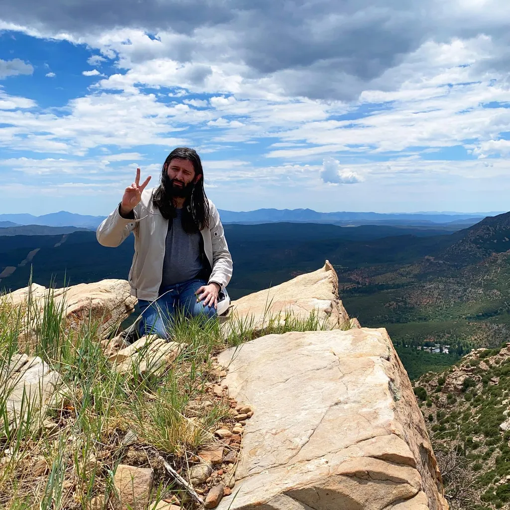 A bearded man in a light jacket stands on a textured rock edge, facing a sweeping view of blue mountains and green valleys beneath a dramatic sky filled with white clouds.