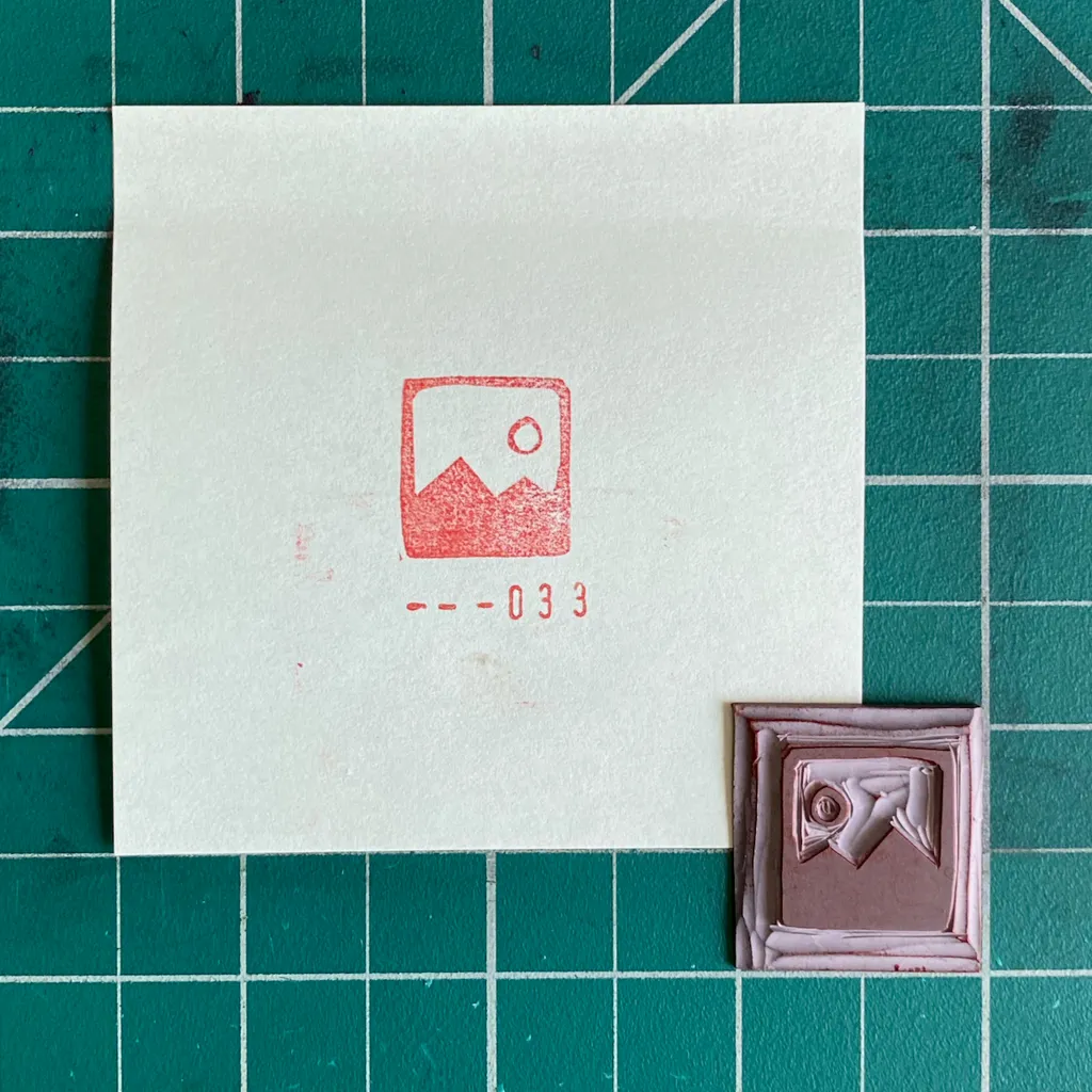 A white card on a green grid displays a red drawing of mountains and a sun with the numbers 033, next to a small, reddish-brown square featuring a raised, stylized figure.