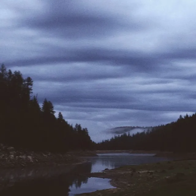 A wide, horizontal landscape shows dark, dense trees framing a calm, reflective river under a heavy expanse of layered gray and blue clouds.