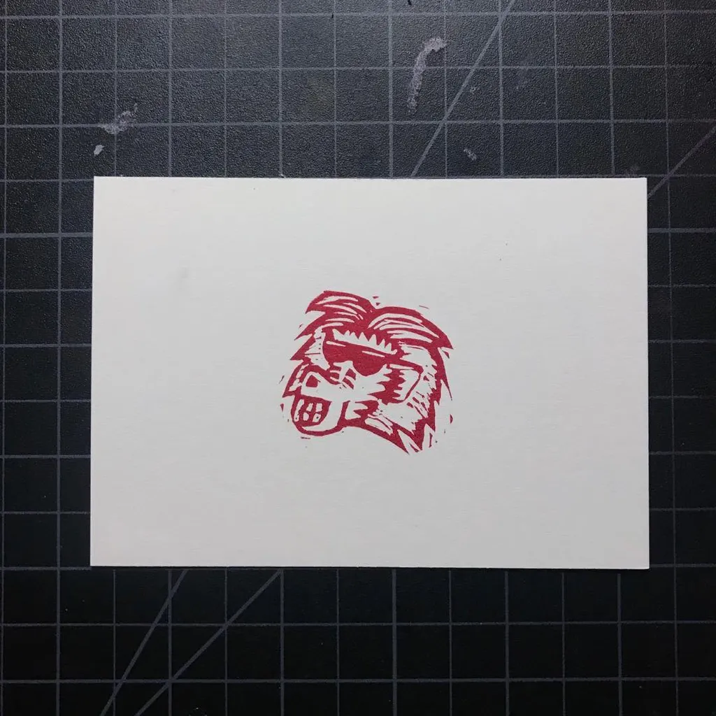 A stylized mask drawing in reddish-pink, executed with rough brushstrokes, is centered on a white paper placed over a dark gray grid background.