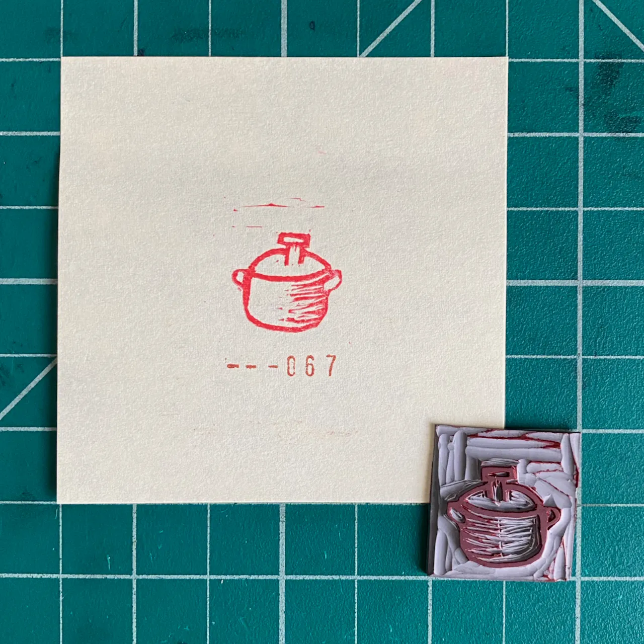 On a teal grid, a cream paper displays a red line drawing of a pot and the number --067, next to a small, textured square artwork featuring a red pot design.
