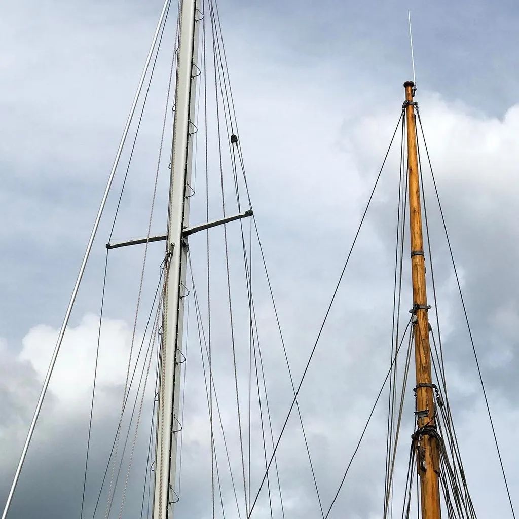 A tall, silver mast stands beside a warm, wooden mast, crisscrossed by many dark rigging lines against a dramatic, cloudy gray sky.