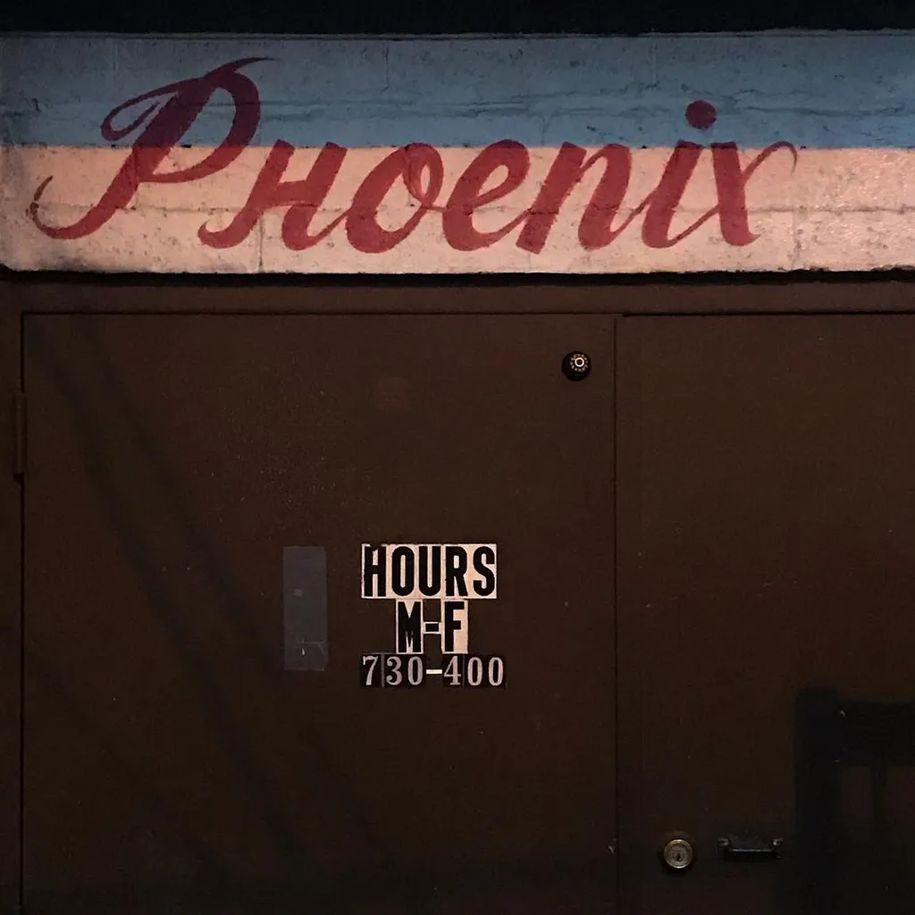 Red script text spelling Phoenix is painted on a light blue wall above a dark brown door that displays white stenciled hours: M-F 7:30-4:00.