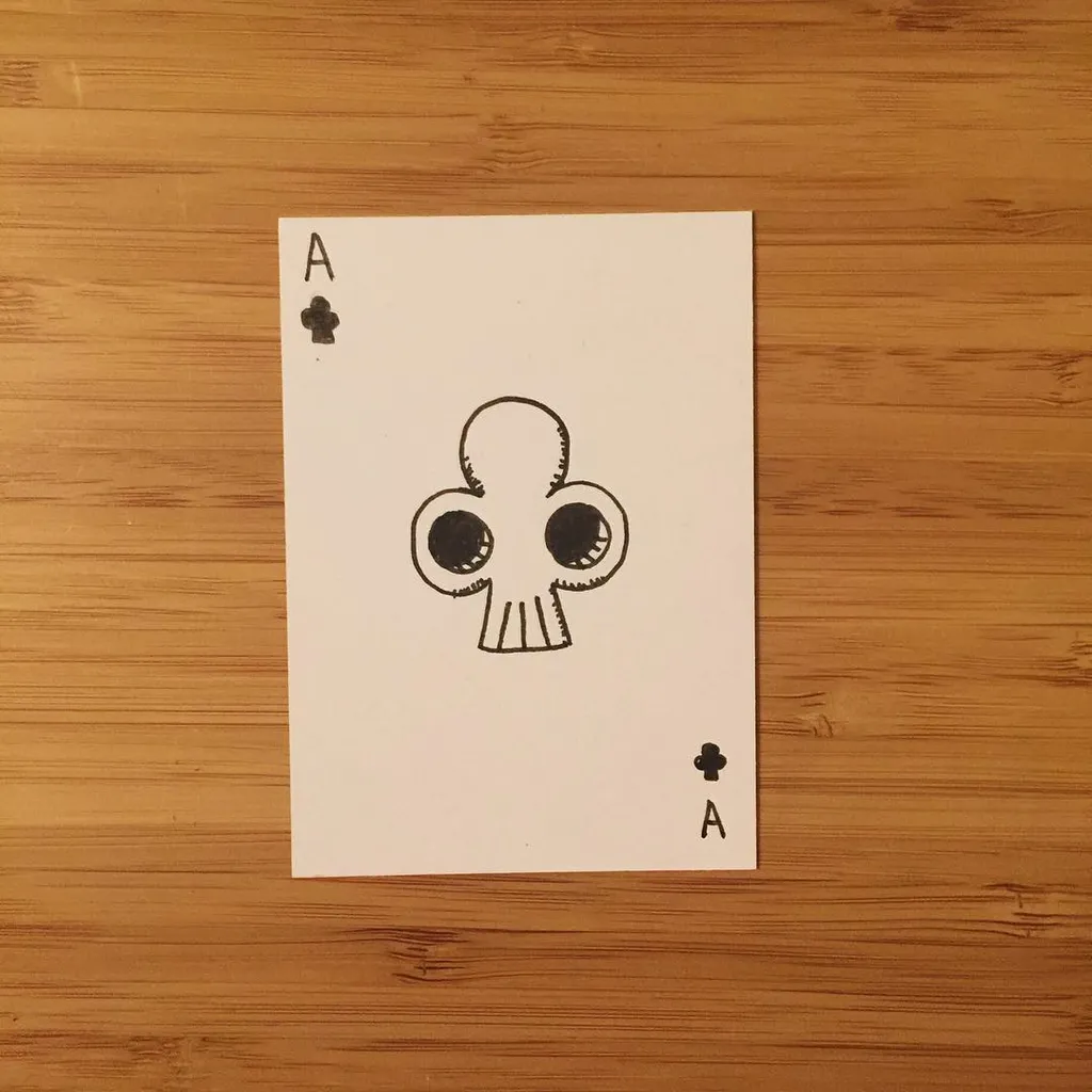 A white playing card featuring an Ace of Clubs, centered on a wooden surface, displays a black line drawing of a stylized, cartoonish face with large eyes and a jaw.