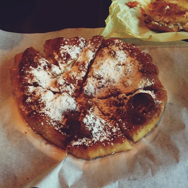 A round, golden-brown baked pastry, dusted with white powder, is cut into wedges and placed on wrinkled light paper, with a second pastry visible in the upper right corner.