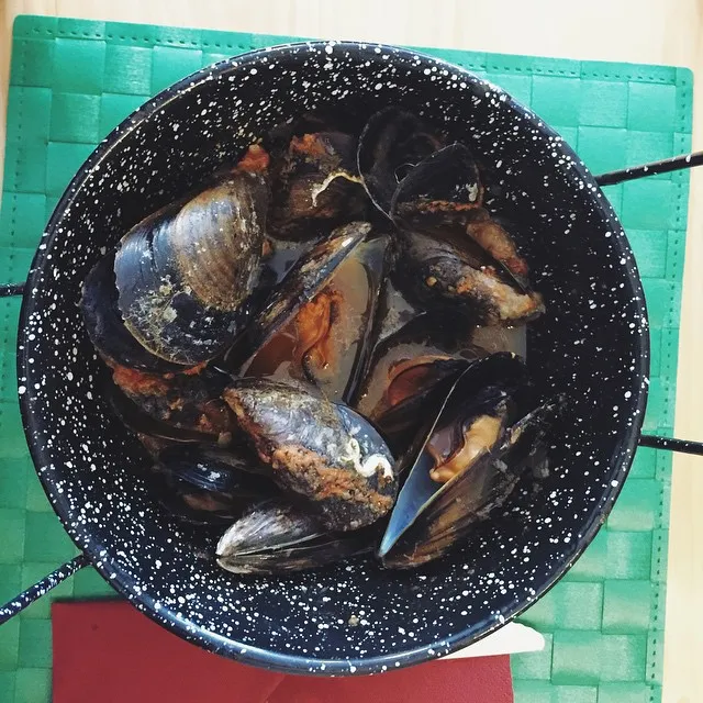 A speckled black pan filled with clustered, dark mussels rests on a bright green mat, with a sliver of red visible beneath the pan.