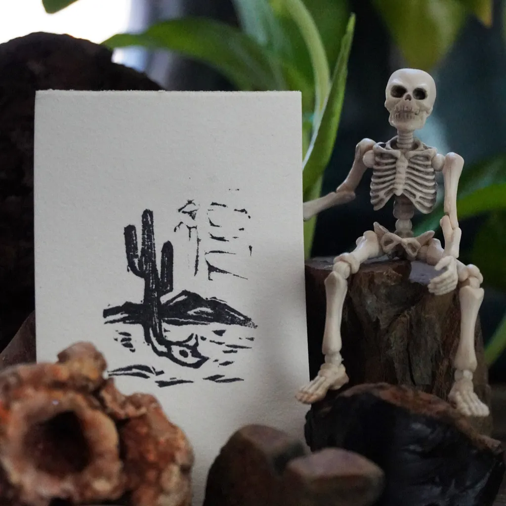 A white card with a black cactus drawing rests beside a white skeleton figurine sitting on dark wood, framed by dark rocks and green foliage.