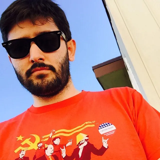 A man with dark hair and beard wears black sunglasses and a red t-shirt featuring a graphic print of figures and symbols, set against a bright blue sky and white building structure.