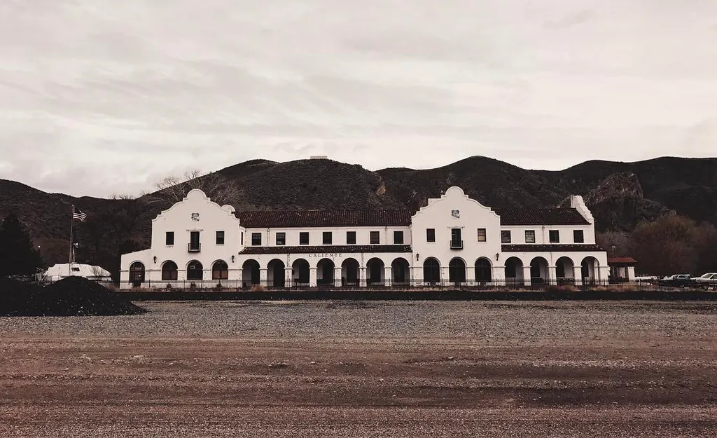 A long, low, white building with arched windows spans the center, set against dark, rolling hills and a wide expanse of textured, gravelly ground under a pale, overcast sky.