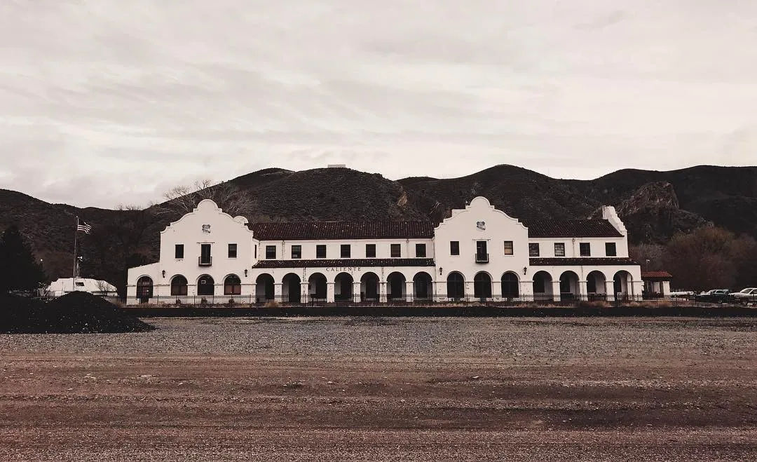 A long, low, white building with arched windows spans the center, set against dark, rolling hills and a wide expanse of textured, gravelly ground under a pale, overcast sky.