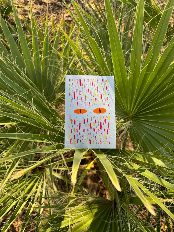 A small white rectangle featuring two orange eyes and vertical drips of yellow, red, and orange paint rests among lush, bright green palm fronds.