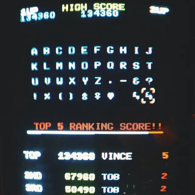 A dark screen displays glowing green text in a structured format, showing high scores, an alphabet key, and a top ranking list with names and numerical scores.