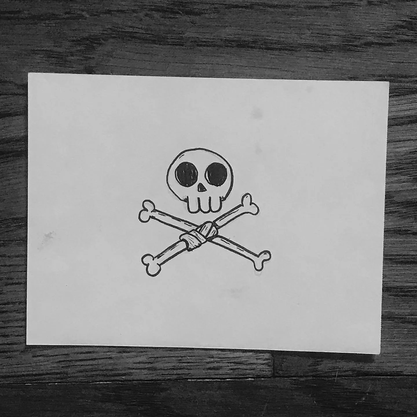 Simple black line art depicts a skull and two crossed bones, drawn on white paper set against a dark wood background.
