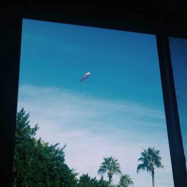 A dark frame surrounds a bright blue sky featuring a small red and white balloon floating above a horizontal line of lush green trees and palm fronds.