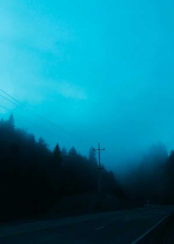 A misty, blue-toned landscape features a dark forest and a road under a bright cyan sky, with a utility pole visible in the center.