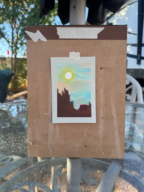 A small, vertical artwork depicting a yellow sun over dark brown silhouettes against a light blue sky is mounted on a larger, plain brown board with visible tape and paper scraps.