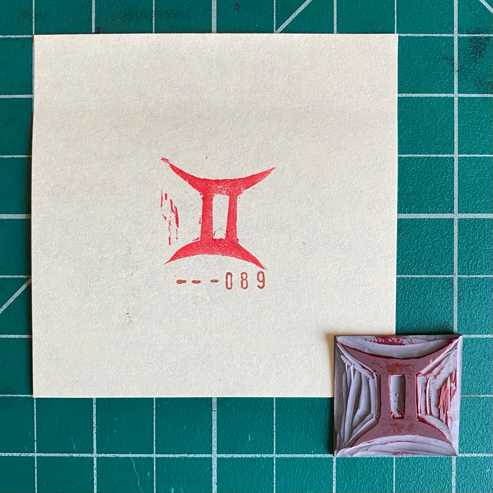A cream paper on a teal grid displays a red stylized graphic and the numbers ---089, next to a small, carved red block that repeats the graphic and numbering.