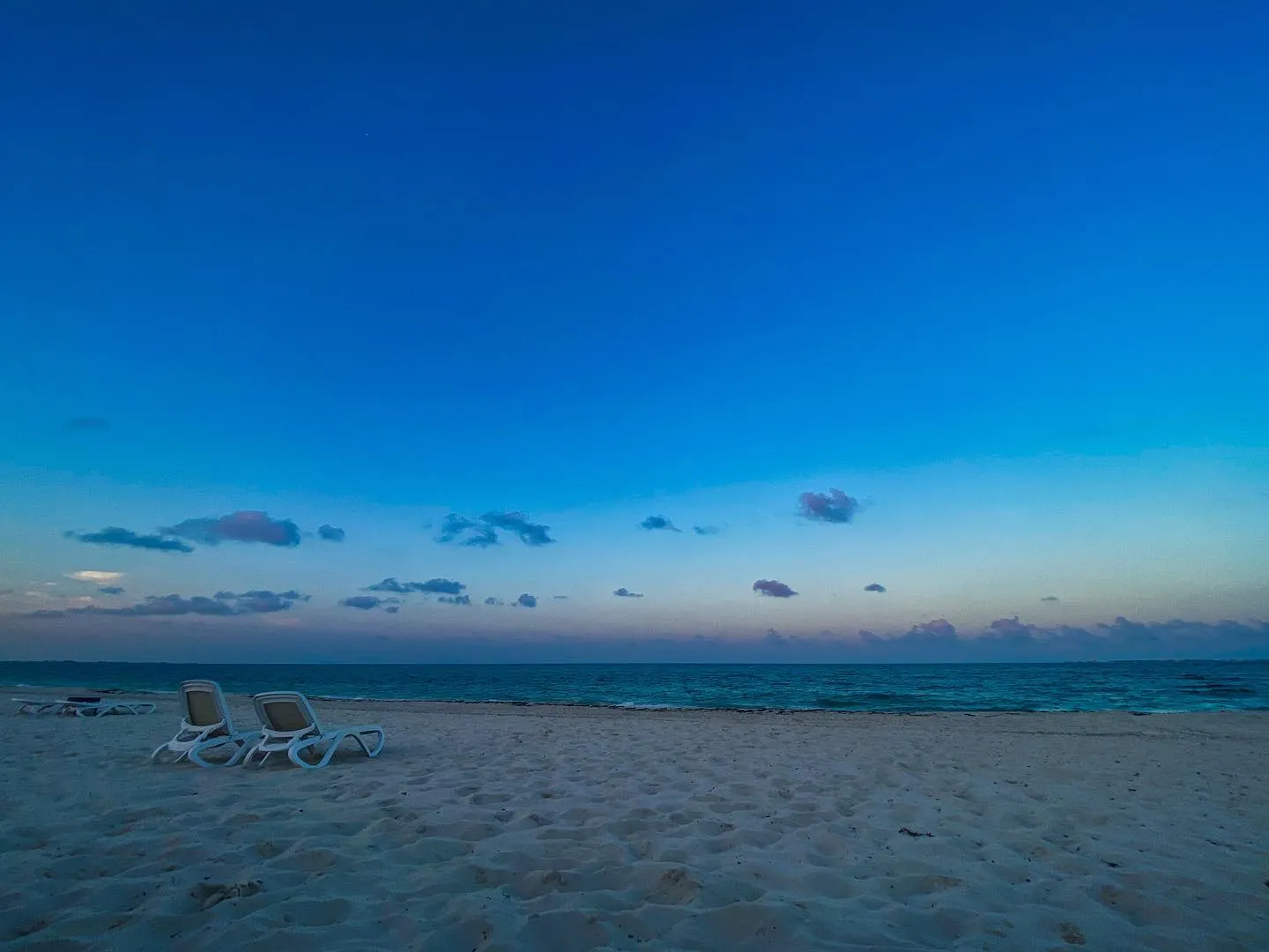 The composition features a vast expanse of deep blue sky and pale sand, meeting a calm blue ocean horizon, with scattered clouds visible across the expanse.