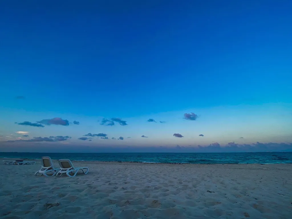 The composition features a vast expanse of deep blue sky and pale sand, meeting a calm blue ocean horizon, with scattered clouds visible across the expanse.